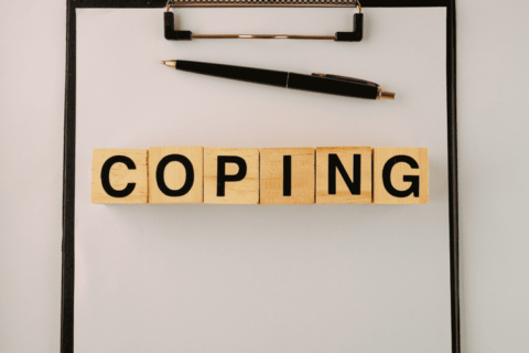 A clipboard with a pen & wooden game tiles spelling the word "Coping"