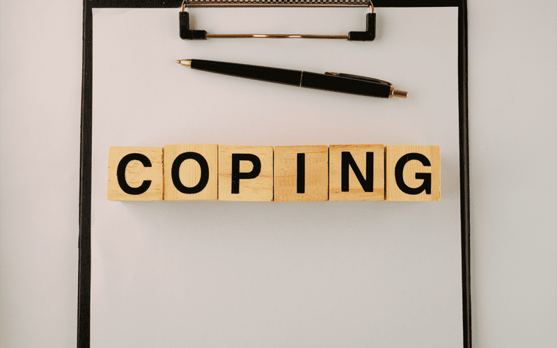 A clipboard with a pen & wooden game tiles spelling the word "Coping"